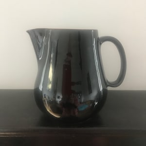 Puede incluir: Una jarra de cerámica negra con cuerpo redondeado y asa. La jarra tiene un acabado brillante.