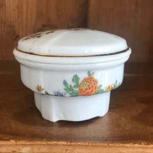May include: A white porcelain trinket box with a floral design. The lid has a gold trim and the box has a floral design with orange, yellow, and green flowers.