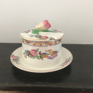 May include: A white porcelain sugar bowl with a floral design and a pink and white radish shaped lid. The bowl has a gold rim and sits on a matching saucer.
