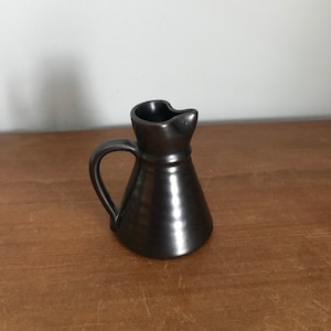 Puede incluir: Un pequeño creamer de cerámica negra con asa. El creamer tiene un cuerpo redondeado y un pico.