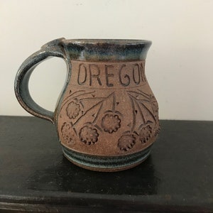 May include: A brown ceramic mug with a blue glaze. The mug has a handle and is decorated with the word "OREGON" and a design of berries.