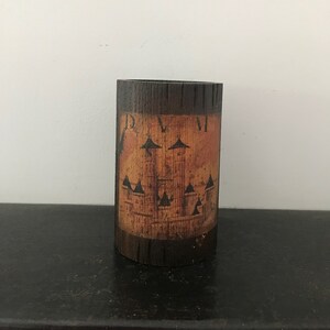 May include: A wooden cylinder with a brown and orange painted design. The design features a castle with three towers and the text "D.V.M."