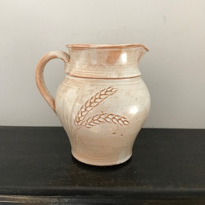 Könnte beinhalten: Ein weißer Keramik Krug mit brauner Glasur und einem Weizenmuster. Der Krug hat einen Henkel und einen runden Körper.