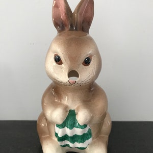 Puede incluir: Una figurita de conejo de cerámica con pelaje marrón, orejas rosas y ojos negros. El conejo sostiene un huevo a rayas verdes y blancas en sus patas.