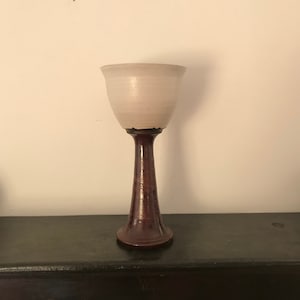 Puede incluir: Una taza de cerámica blanca con una base de madera marrón. La taza tiene una forma redondeada y un borde ancho. La base es alta y delgada.