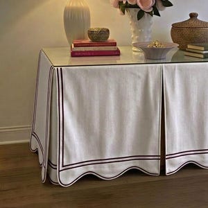 May include: A rectangular console table covered with a white fabric skirt featuring a scalloped hem and dark red trim. The table surface holds a white lamp, a vase of pink flowers, a woven lidded bowl, and two stacks of books under a framed print.