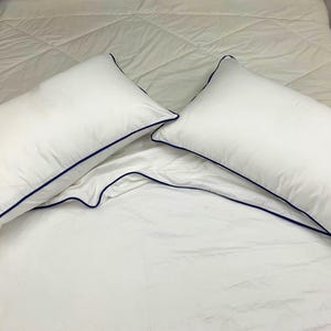 Puede incluir: Dos fundas de almohada blancas con un borde azul oscuro sobre una colcha blanca.