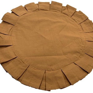 May include: A round, tan-colored placemat with a pleated ruffle border. The placemat is made of a soft fabric and is designed to protect a table from spills and heat. The overall design is simple and elegant.