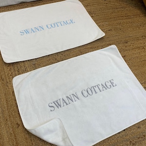 Puede incluir: Dos fundas de almohada rectangulares blancas con el texto "SWANN COTTAGE" impreso en azul y gris. Las fundas de almohada están sobre una superficie marrón tejida.