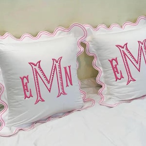 May include: Two white square pillows with pink embroidered initials and scalloped edges. The pillows are on a white bedspread. The embroidered initials are in a decorative font.