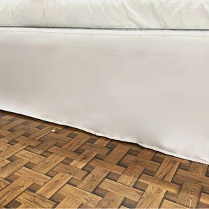 Może przedstawiać: Biała spódnica do łóżka o prostym wzorze, wisząca nad podłogą drewnianą z plecionym wzorem.