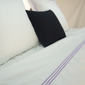 Puede incluir: Bedding blanco con tres rayas moradas que recorren el centro de la s&aacute;bana. Dos almohadas blancas con ribetes morados est&aacute;n en la cama, y una almohada negra est&aacute; encima de ellas.