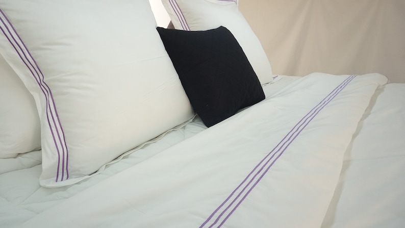 Puede incluir: Bedding blanco con tres rayas moradas que recorren el centro de la s&aacute;bana. Dos almohadas blancas con ribetes morados est&aacute;n en la cama, y una almohada negra est&aacute; encima de ellas.