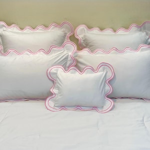 Puede incluir: Un juego de fundas de almohada blancas con un ribete rosa festoneado. El juego de cama incluye almohadas rectangulares de varios tamaños, dispuestas sobre una colcha blanca. El ribete rosa añade un toque decorativo a las almohadas blancas.
