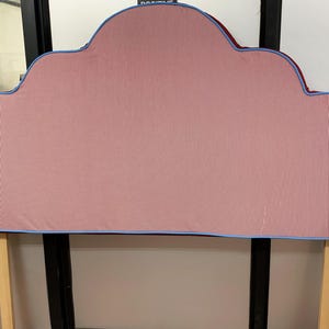 Pode incluir: Uma cabeceira em forma de nuvem com um padrão de riscas rosa e branco. A cabeceira tem uma borda azul e é suportada por duas pernas de madeira. O design é suave e convidativo, adequado para um quarto. Mede aproximadamente 150 cm de largura.