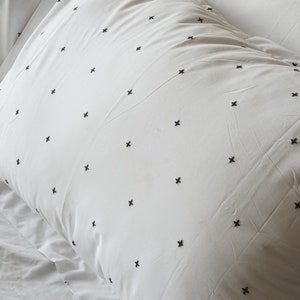 May include: White pillowcases with a repeating pattern of small black cross-stitch embroidery. The pillowcases are on a white bedsheet, creating a clean and minimalist aesthetic. The bedding appears soft and comfortable.