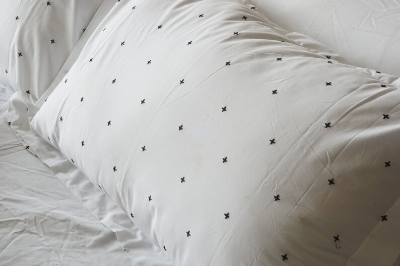 May include: White pillowcases with a repeating pattern of small black cross-stitch embroidery. The pillowcases are on a white bedsheet, creating a clean and minimalist aesthetic. The bedding appears soft and comfortable.