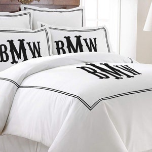 K&ouml;nnte beinhalten: Wei&szlig;es Bettw&auml;sche-Set mit schwarzen Monogramm-Initialen und schwarzem Rand. Das Set beinhaltet Kissenbez&uuml;ge und einen Bettbezug. Die Initialen sind gro&szlig; und mittig auf der Bettdecke und den Kissenbez&uuml;gen platziert.