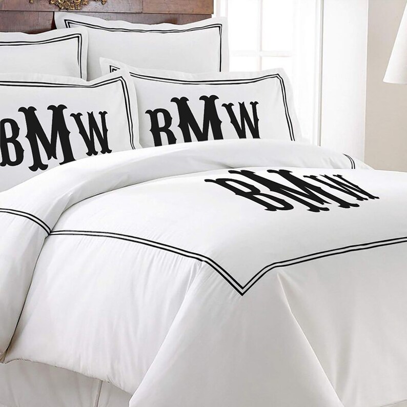 K&ouml;nnte beinhalten: Wei&szlig;es Bettw&auml;sche-Set mit schwarzen Monogramm-Initialen und schwarzem Rand. Das Set beinhaltet Kissenbez&uuml;ge und einen Bettbezug. Die Initialen sind gro&szlig; und mittig auf der Bettdecke und den Kissenbez&uuml;gen platziert.