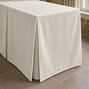 May include: A rectangular table cover in a solid light beige color with a textured fabric finish. The cover features a structured design with sharp, clean corners and vertical pleats along the sides, accented by crisp white piping trim.