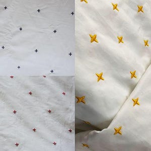 May include: White fabric featuring embroidered cross and star patterns. The crosses are navy and red, the stars are gold. The material appears to be cotton or linen, suitable for clothing or home decor.