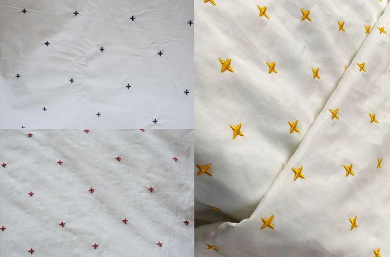 May include: White fabric featuring embroidered cross and star patterns. The crosses are navy and red, the stars are gold. The material appears to be cotton or linen, suitable for clothing or home decor.