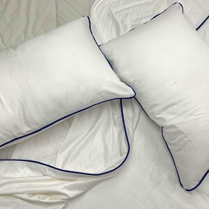 Puede incluir: Dos fundas de almohada blancas con un ribete azul oscuro. Las fundas de almohada est&aacute;n sobre una s&aacute;bana de cama blanca.