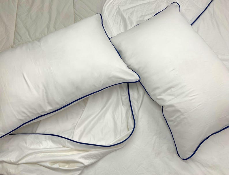 Puede incluir: Dos fundas de almohada blancas con un ribete azul oscuro. Las fundas de almohada est&aacute;n sobre una s&aacute;bana de cama blanca.