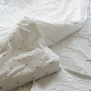 May include: White bedding set with a minimalist design. The duvet cover and pillowcases feature a pattern of small black crosses. The sheets are slightly rumpled, suggesting a comfortable and inviting aesthetic.