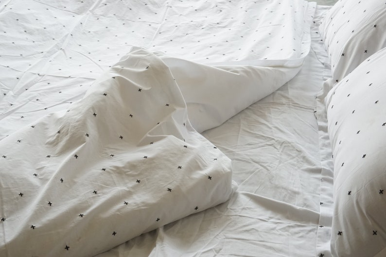 May include: White bedding set with a minimalist design. The duvet cover and pillowcases feature a pattern of small black crosses. The sheets are slightly rumpled, suggesting a comfortable and inviting aesthetic.