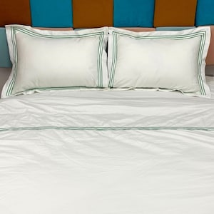 Puede incluir: S&aacute;banas blancas de cama con un borde bordado verde. Las s&aacute;banas est&aacute;n en una cama con una cabecera hecha de tela marr&oacute;n, azul y rosa.