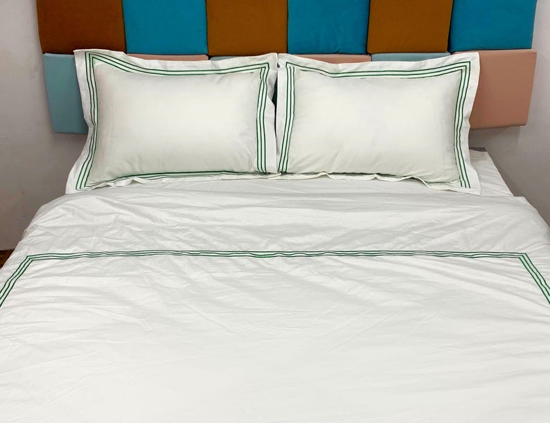 Puede incluir: S&aacute;banas blancas de cama con un borde bordado verde. Las s&aacute;banas est&aacute;n en una cama con una cabecera hecha de tela marr&oacute;n, azul y rosa.