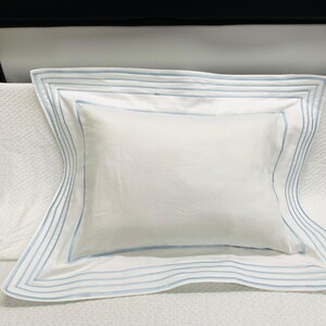 Puede incluir: Una almohada blanca con ribete azul. El ribete es una serie de líneas finas alrededor del borde de la almohada.