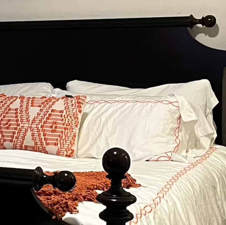 Puede incluir: Una cabecera de cama de madera negra con una almohada estampada en blanco y rojo en la parte superior. La cama est&aacute; hecha con s&aacute;banas blancas y un edred&oacute;n blanco con ribete rojo.