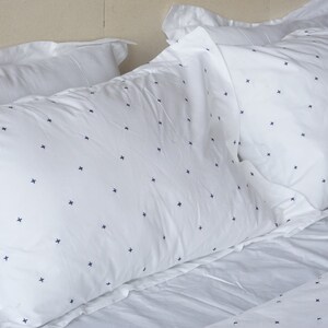 May include: White bed linen set featuring a navy blue cross pattern. The image displays several pillows and a sheet, all with the same design. The pillows are arranged on a bed, creating a clean and simple aesthetic.