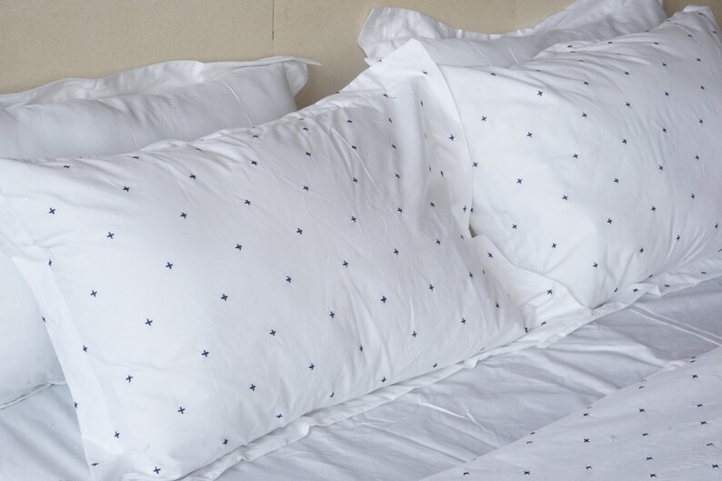 May include: White bed linen set featuring a navy blue cross pattern. The image displays several pillows and a sheet, all with the same design. The pillows are arranged on a bed, creating a clean and simple aesthetic.