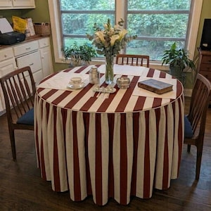 May include: A round dining table covered with a floor-length, pleated tablecloth featuring vertical stripes in dark red and off-white. The table is set with a white teacup, a small glass jar, a silver spoon, and a rectangular brown book.
