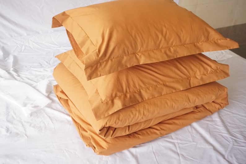May include: A stack of folded mustard yellow bedding, including a duvet and pillowcases, on a white surface.