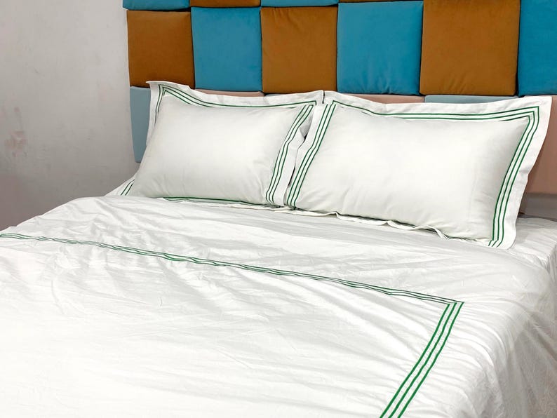 Puede incluir: Ropa de cama blanca con ribete verde. La ropa de cama est&aacute; en una cama con una cabecera hecha de cuadrados marrones y azules.
