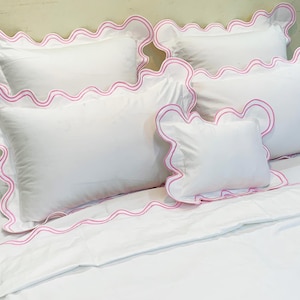 Puede incluir: Juego de cama blanco con ribete festoneado rosa. El juego incluye varias almohadas de diferentes tamaños y una sábana bajera. Las almohadas están dispuestas sobre una cama, mostrando el ribete decorativo.