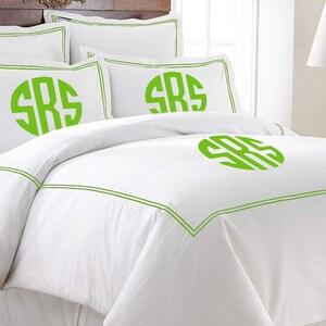 K&ouml;nnte beinhalten: Wei&szlig;es Bettw&auml;sche-Set mit gr&uuml;nen Zierleisten und Monogramm. Das Set beinhaltet einen Bettbezug und Kissenbez&uuml;ge, alle mit den Initialen "SRS" in leuchtendem Gr&uuml;n. Die Bettw&auml;sche hat ein sauberes, frisches Aussehen.