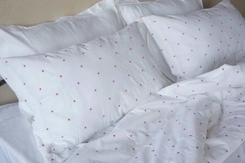 May include: White bedding set featuring red cross-stitch embroidery. The set includes two pillowcases and a duvet cover, all made from soft, white fabric. The red crosses are evenly spaced, creating a simple, elegant design. The duvet cover and pillows are on a bed.