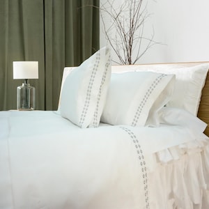 May include: White bed linens with a ruffled edge and a subtle gray embroidered pattern. The bed is made with two pillows, one with a gray embroidered pattern and one with a plain white cover.
