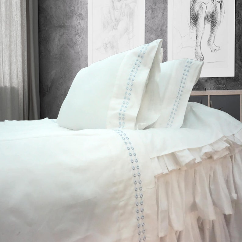May include: White bed linens with a ruffled edge and a blue embroidered design. The linens are made of a soft, comfortable fabric and are perfect for a bedroom.