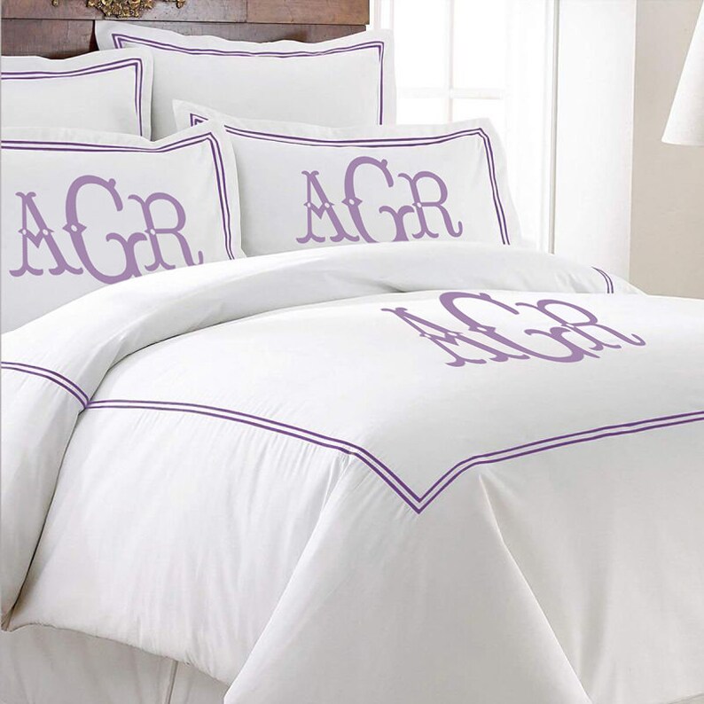 K&ouml;nnte beinhalten: Wei&szlig;es Bettw&auml;sche-Set mit lila Zierleisten und Monogramm-Initialen. Das Set beinhaltet Kissenbez&uuml;ge und einen Bettbezug. Die Initialen "AGR" sind auf den Kissenbez&uuml;gen und dem Bettbezug zu sehen und verleihen dem Schlafzimmerdekor eine pers&ouml;nliche Note.