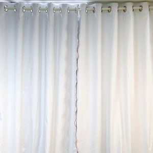 May include: White curtains with a scalloped red trim. The curtains are hanging from a silver rod with grommets. The fabric appears to be a light, semi-sheer material, creating a soft, flowing look. The curtains are likely used for window coverings.