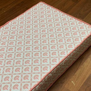 Op de afbeelding: Rechthoekig kussen met een herhalend bloemmotief in lichtroze op een witte achtergrond. Het kussen heeft een roze rand. Waarschijnlijk voor woondecoratie of meubels.