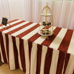 May include: A rectangular table covered with a fabric cloth featuring vertical stripes in dark red and off-white. On the table sits a gold-toned metal three-tiered serving stand holding assorted chocolates and a small black sign labeled Candy.