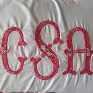K&ouml;nnte beinhalten: Wei&szlig;es Kissen mit gestickten rosa Buchstaben, die "CSA" buchstabieren. Die Buchstaben haben dekorative Verzierungen und sind in einem kr&auml;ftigen, kursiven Stil gestickt. Das Kissen hat einen satinartigen Glanz.