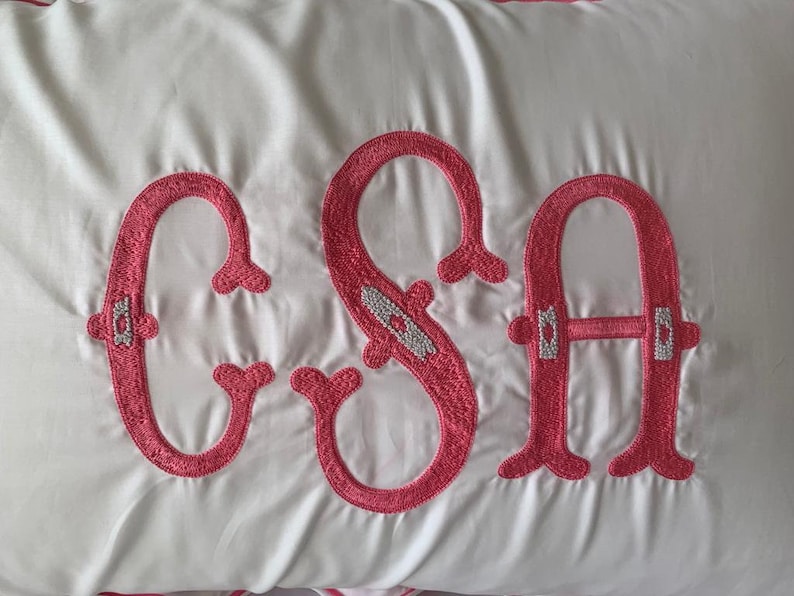 K&ouml;nnte beinhalten: Wei&szlig;es Kissen mit gestickten rosa Buchstaben, die "CSA" buchstabieren. Die Buchstaben haben dekorative Verzierungen und sind in einem kr&auml;ftigen, kursiven Stil gestickt. Das Kissen hat einen satinartigen Glanz.